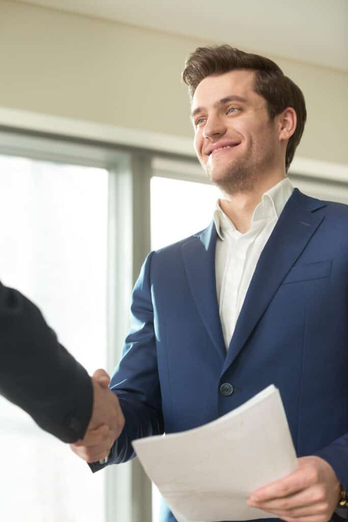 Confident smiling businessman with documents in hand handshaking with partner in office