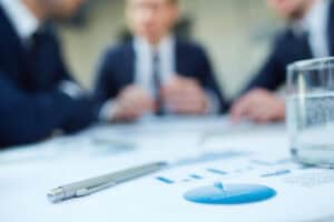 Image of business document and pen at workplace with group of colleagues interacting in background