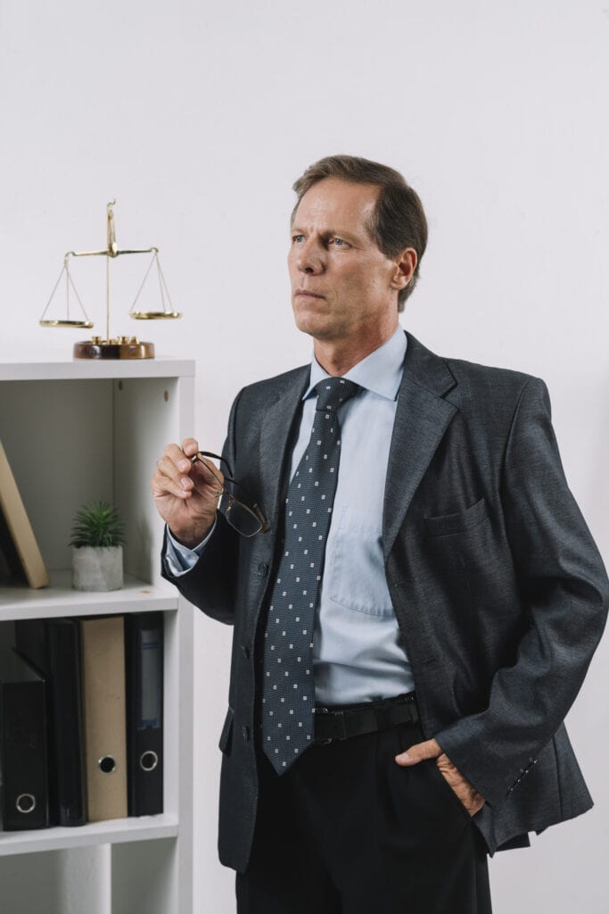 Man in suit standng in office with hand in his pocket, scale of justice in the backround