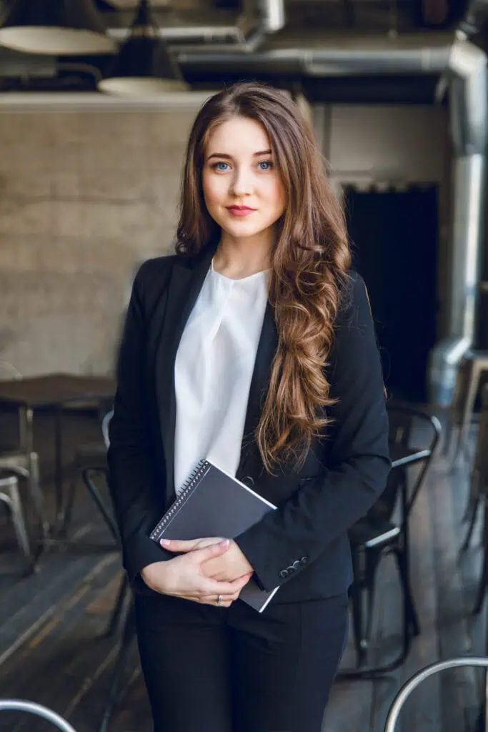 Brunette business woman in a suit with wavy long hair and blue eyes stands holding a notebook in hands