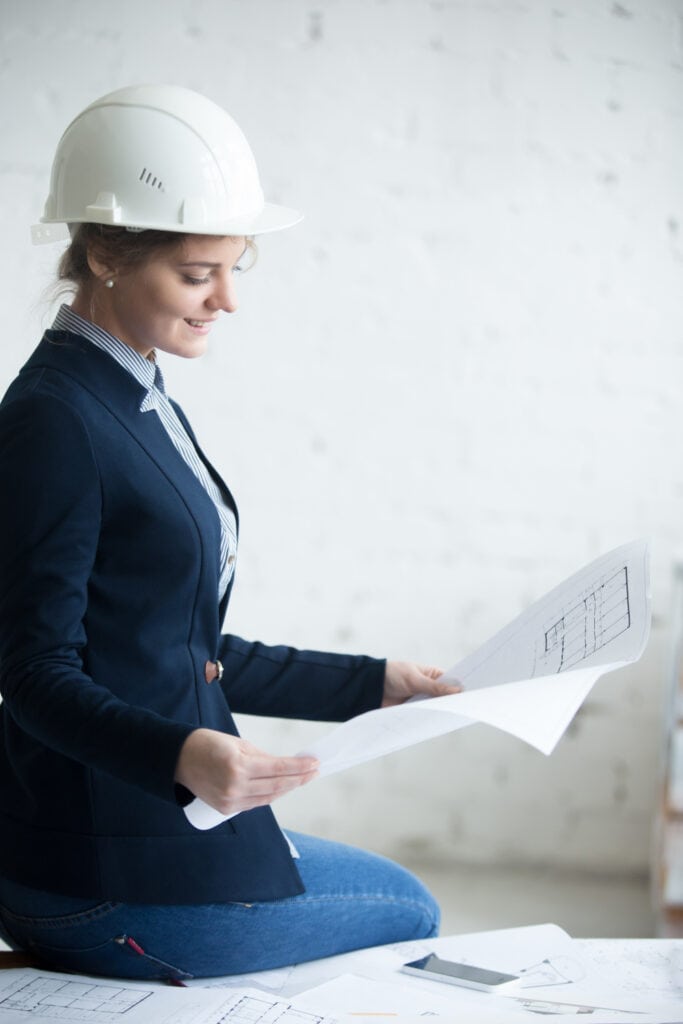 Female SQA engineer wearing a hard white helmet is sitting on a table, holding a project plan in her hands