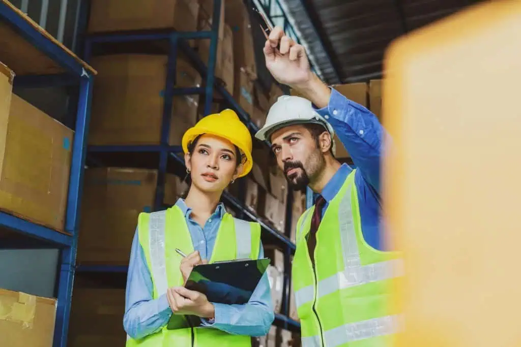 Two logistic experts wearing reflexive vest and hard helmets stand in warehouse, with the man pointing out and the woman taking notes