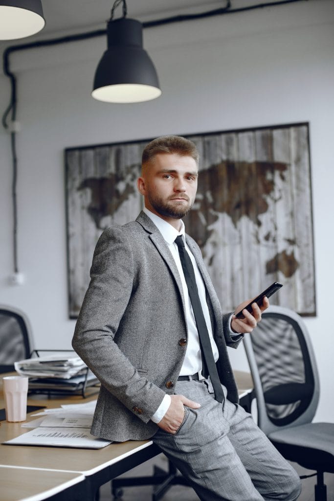 Businessman in grey suit holding a phone is leaning on his desk in an office